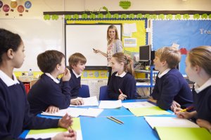 pupils sitting at table as teacher stands by whiteboard